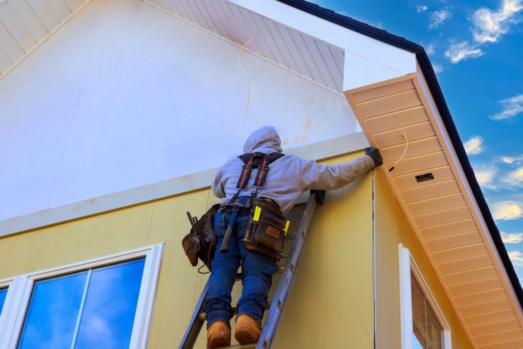 Worker is using ladder to fix trim on house while installing fiber siding panels